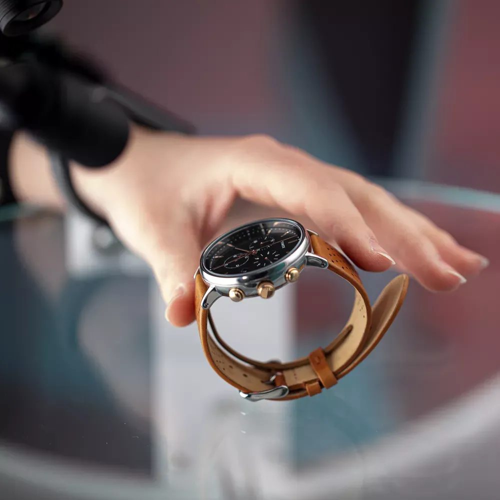 Hand positioniert eine Herrenuhr mit braunem Lederarmband auf einer Glasfläche